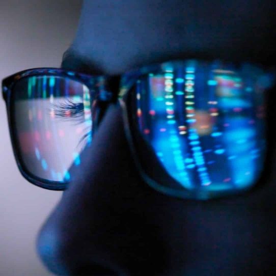 Closeup image of a man’s glasses reflecting multicolored data visualization on a computer screen